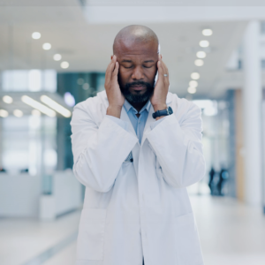 Overworked doctor rubbing his temples in a busy hospital, showing signs of stress and burnout, with blurred medical staff rushing in the background."